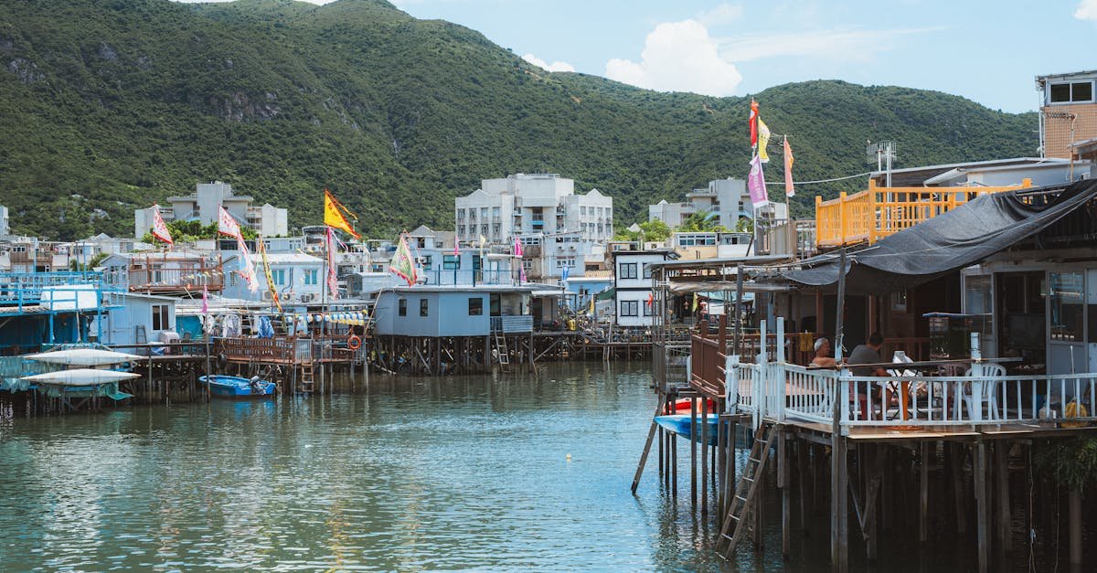 Kuće na stupovima sa šarenim zastavama stoje nad mirnom vodom u selu okruženom zelenim brdima. Neke zgrade imaju vanjska mjesta za sjedenje, a u blizini su privezani brodovi. Nebo je djelomično oblačno, stvarajući spokojnu atmosferu.