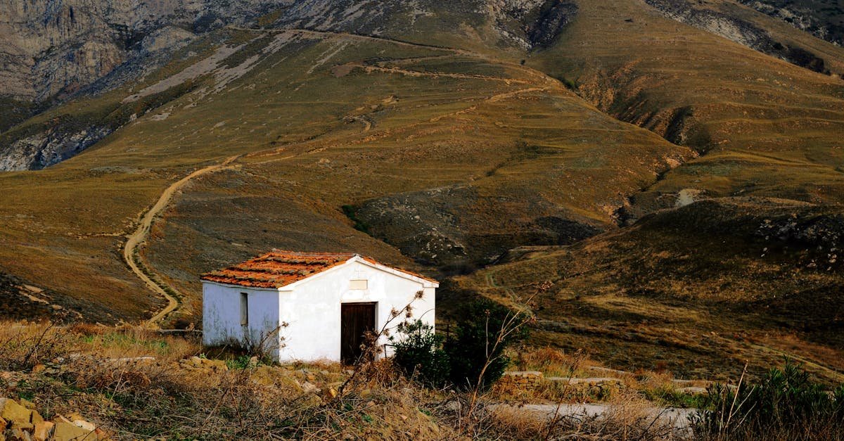 Mala bijela kuća s krovom od crvenog crijepa stoji sama na suhom, travnatom obronku. Vijugave zemljane staze vijugaju kroz surova smeđa brda i planine u pozadini pod vedrim nebom. Rijetka vegetacija okružuje izoliranu kuću.