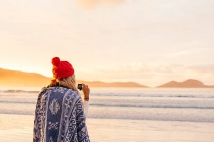 Osoba omotana plavim uzorkom dekice i crvenom kapom stoji na plaži pri izlasku ili zalasku sunca, držeći fotoaparat i okrenuta prema oceanu, s planinama i mekim zlatnim svjetlom u pozadini.