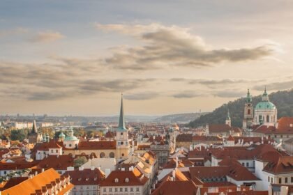 Panoramski pogled na europski grad s krovovima od crvenog crijepa, tornjevima i crkvama s kupolama pod djelomično oblačnim nebom u zalazak sunca. Na horizontu se vide brda i udaljene moderne zgrade, stvarajući kontrast između stare i nove arhitekture.