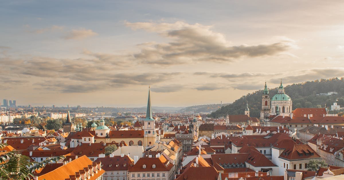 Panoramski pogled na europski grad s krovovima od crvenog crijepa, tornjevima i crkvama s kupolama pod djelomično oblačnim nebom u zalazak sunca. Na horizontu se vide brda i udaljene moderne zgrade, stvarajući kontrast između stare i nove arhitekture.
