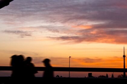 Siluete figura hodaju preko mosta u zalazak sunca, s narančastim i ljubičastim nebom. Na horizontu se vide TV toranj i udaljene zgrade. U sceni se nalaze i horizontalne ograde i ulična svjetiljka.