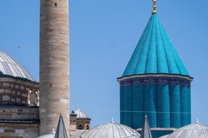 Slika prikazuje dio džamije s visokim bež minaretom, kupolama na krovovima i stožastom kupolom od tirkiznih pločica sa zlatnim ukrasima naspram vedrog plavog neba. Arhitektura prikazuje bliskoistočne i islamske elemente dizajna.