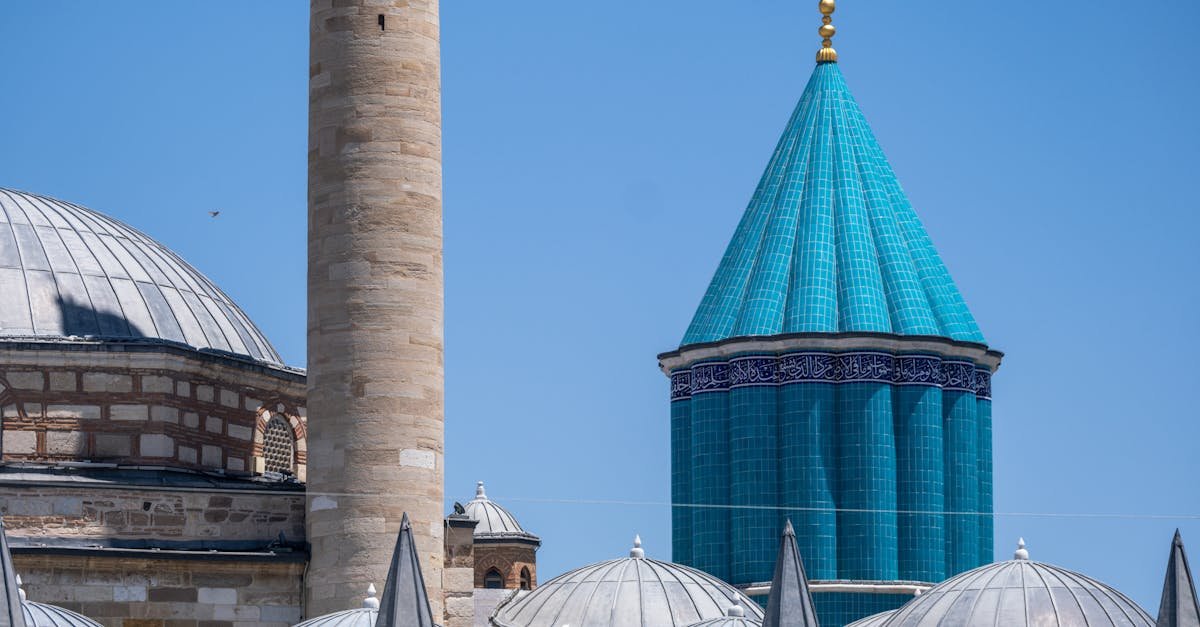 Slika prikazuje dio džamije s visokim bež minaretom, kupolama na krovovima i stožastom kupolom od tirkiznih pločica sa zlatnim ukrasima naspram vedrog plavog neba. Arhitektura prikazuje bliskoistočne i islamske elemente dizajna.
