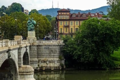 Fotografija kamenog mosta s ukrašenim kipovima koji prelaze mirnu rijeku, okružen drvećem i povijesnim, šarenim zgradama. U pozadini su vidljiva zelena brda i oblačno nebo, stvarajući spokojni europski gradski pejzaž.