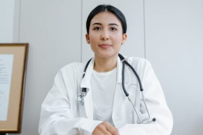 Liječnica s tamnom kosom svezanom unatrag, odjevena u bijeli mantil i sa stetoskopom, sjedi za stolom. Dijagram ljudskog mišićnog sustava uokviren je na zidu iza nje. Gleda naprijed s neutralnim izrazom lica u kliničkom okruženju ordinacije.