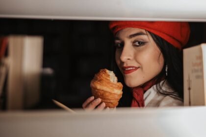Žena u crvenoj beretki i šalu smiješi se dok drži kroasan. Stoji između polica s knjigama, s knjigama vidljivim s obje strane, i gleda u kameru u mekom okruženju.