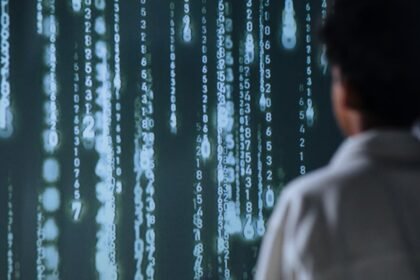 Osoba s kratkom, kovrčavom kosom, gledana straga, u slabo osvijetljenom okruženju, na velikom digitalnom zaslonu koji prikazuje okomite redove svjetlećih brojeva i simbola, nalik zelenom kodu iz Matrice.