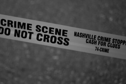 Crno-bijela fotografija prikazuje policijsku vrpcu s natpisima MJESTO ZLOČINA ZABRANJEN PRELAZAK i NASHVILLE CRIME STOPPERS NOVAC ZA TRAGOVE 74-ZLOČIN razapetu preko zamućene pozadine, što ukazuje na ograničeno područje istrage.