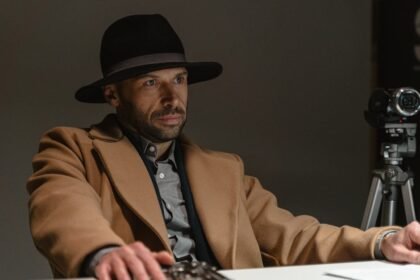 Ozbiljan muškarac u crnom šeširu širokog oboda i smeđem kaputu sjedi za stolom prekriženih ruku. Okrenut je prema naprijed pod dramatičnim osvjetljenjem. Na stolu su papiri i naočale. Videokamera na tronošcu postavljena je iza njega. Pozadina je tamna.