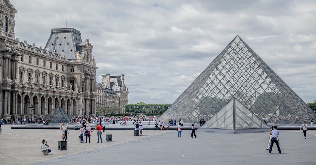 Ljudi šetaju dvorištem muzeja Louvre u Parizu, gdje se pod oblačnim nebom vide veliki ulaz u staklenu piramidu, dvije manje staklene piramide i povijesna kamena zgrada muzeja.