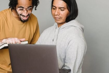 Dvoje ljudi stoje blizu jedno drugome i gledaju u laptop. Jedna osoba nosi smeđu majicu i drži knjigu, oduševljeno se smiješeći. Druga osoba, u sivoj majici s kapuljačom, drži laptop i izgleda usredotočeno. Pozadina je jednostavna i neutralna.