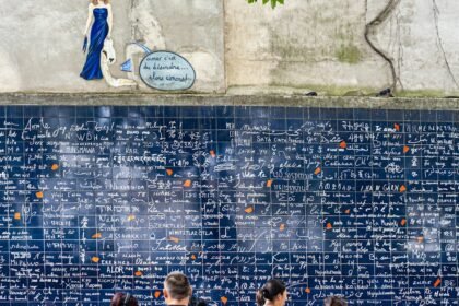 Grupa ljudi stoji ispred velikog popločanog zida prekrivenog natpisom "Volim te" na raznim jezicima. Zid i natpisi su uglavnom plavo-bijeli. Iznad zida nalazi se mali mural žene u plavom i bijelog balona s tekstom.